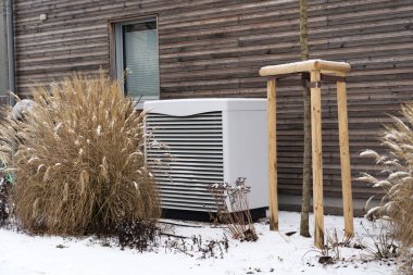 Modern ahşap bir duvarın yanına kurulmuş enerji tasarruflu ısı pompası. Ünitenin etrafı kar katmanlı kuru otlarla çevrili. Kış manzarasında çevre dostu ısıtma teknolojisi sergiliyor..