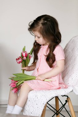 Parlak bir odada bir sandalyede oturan ve bir buket taze pembe laleyle oynayan küçük bir çocuk. Aile hayatı, çocuklar ve mevsimsel içerik için ideal yumuşak ışıklı basit bir kompozisyon..