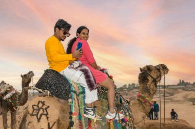 İki kişi çölde gün batımında deve sürüşünden zevk alır. Hindistan veya Rajasthan 'da seyahat, turizm, yaşam tarzı, macera ve kültür hikayeleri temaları için canlı giysiler, püsküller ve manzaralı kayakların yer aldığı.