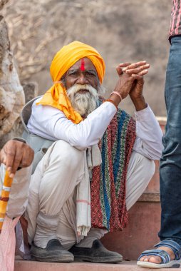 Yaşlı Hintli bir adam, parlak sarı bir türban, beyaz bir elbise ve renkli bir kumaş giyerek taş basamağa çömeliyor. Kırmızı bir tilak alnını işaret ediyor. Yaşam tarzı, seyahat ve portre temaları için kültürel derinlik ve ruhani varlıkları yakalar.