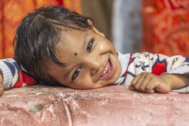 Islak saçlı, renkli elbiseli neşeli çocuk pembe bir yüzeye yaslanırken gülümsüyor. Sıcak tonlar ve canlı kumaş arka plan bir Hint ortamında samimi duygular ve kültürel cazibe yakalar..