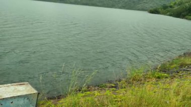Hindistan 'ın Tamil Nadu kentindeki Aliyar Baraj Barajı' nda sakin bir göl manzarası. Çimen kıyısında paslı metal bir kutu, hala berrak su ve uzakta da hafif bir yamaç var. Huzurlu doğal atmosfer