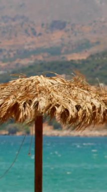 Barışçıl bir gündüz manzarasında sahilde bir kamış şemsiyesi, şeffaf mavi suyun yavaşça kıyıya vurduğu görülüyor. Yeşil tepeler güneşli gökyüzünün altında manzaralı bir zemin sağlar..