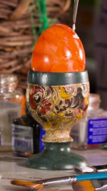 Bir sanatçı canlı turuncu bir yumurtayı dekoratif bir tutacağa dikkatlice resmeder. Çalışma alanını çevreleyen çeşitli sanat malzemeleri yaratıcılık sürecini gözler önüne seriyor..