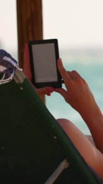 Plaj sandalyesinde, güneşin altında bir e-okuyucu okurken huzurlu bir anın tadını çıkar. Okyanus dalgaları bu rahatlatıcı aktivite için rahatlatıcı bir zemin oluşturur..