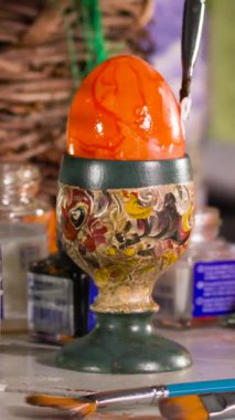 Parlak turuncu boya, nefis bir el yapımı tutacakta dinlenen bir yumurtaya uygulanır. Sanat eserlerinin etrafındaki malzemeler bahar sanatı seansı sırasında yaratıcı bir atmosfere işaret ediyor..