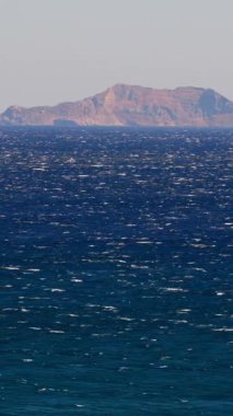 Denizde gün ışığı parıldarken kumsal boyunca dalgalar yavaşça kırılır. Arka planda, sakin bir ada sessizce uzaklarda oturur ve manzaraya güzellik katar..