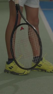 Genç bir tenisçi yeşil bir kortta maç başlatmaya hazır. Oyuncu raketi sıkıca tutarken parlak sarı ayakkabılar göze çarpıyor. Odaklanma ve beklenti açık..