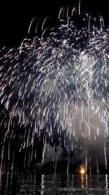 Parlak havai fişekler durgun suyun üzerinde renkli görüntüler oluşturarak seyirciyi büyüleyen bir yaz akşamı etkinliği sırasında şenlikli bir atmosfer yaratıyor..