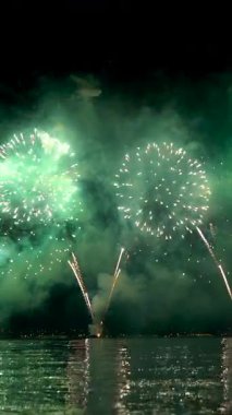 Gece gökyüzünde, dingin bir su kütlesinin üzerinde renkli havai fişekler patlıyor, canlı renkleri yansıtıyor ve yakınlarda toplanan izleyiciler için neşe saçıyor..