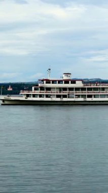 Büyük bir nehir teknesi bulutlu bir gökyüzünün altında durgun suda sorunsuzca süzülür. Yolcular, tekne nehir boyunca ilerlerken manzaranın tadını çıkarıyorlar..