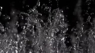Su damlacıkları çarpışıp sıçrıyor ve karanlık bir zemine karşı karmaşık desenler oluşturuyor. Yavaş çekim etkisi, anın güzelliğini ve ışığın yansımalarını vurgular..