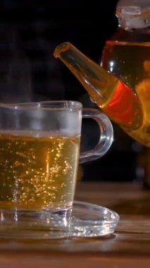 Bir çaydanlık temiz bir bardağı sıcak çayla doldurur, yüzeyden yükselen buharla. Sıcak renkler ve yumuşak ışık rahatlatıcı bir içeceğin keyfini çıkarmak için mükemmel bir ortam yaratır..