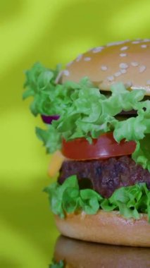 Parlak sarı bir zemin üzerinde taze marul, domates ve peynirle kaplı lezzetli bir hamburgerin yakın plan çekimi, canlı renkleri ve dokuları vurguluyor..