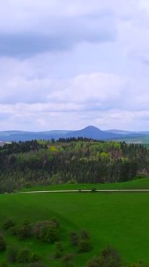 Bereketli yeşil tepelerin ve bulutlu bir gökyüzünün altındaki uzak dağın huzurlu manzarasının tadını çıkar. Manzara, bahar mevsiminde doğanın güzelliğini sergiliyor..