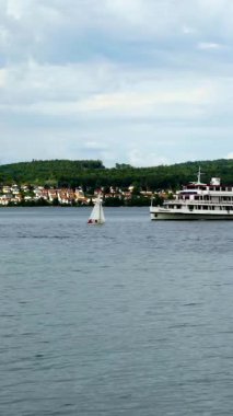 A small sailboat navigates through tranquil waters beside a large ferry on a beautiful lake. Lush green hills frame the backdrop, creating a serene atmosphere.