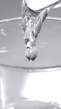 İyi aydınlatılmış bir mutfakta kaşıktan şeffaf bir bardağa düşen temiz bir sıvı damlası görülüyor. Bu an, basit mutfak aktivitelerinin güzelliğini yakalar..