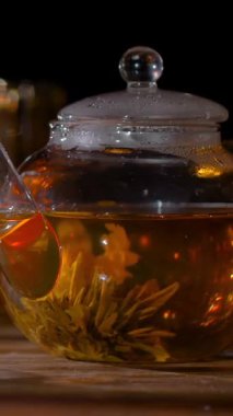 Bir cam çaydanlık masanın üzerinde oturur. Sıcak su saksının içindeki çiçeklerin üzerine dökülüyor. Bir kaşık çayı karıştırır. Tencereden buhar yükseliyor. Bu sessiz bir demlenme anı..