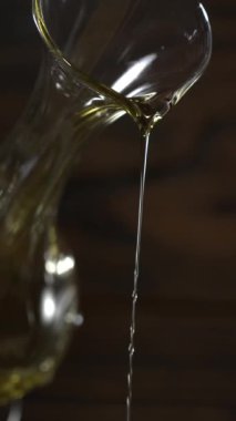 Cam bir kap eğilir ve zeytin yağı ahşap bir masaya akar. Işık hareket ederken sıvının üzerinde parlıyor. Mutfakta doğal ışık ile sıcak bir his var..