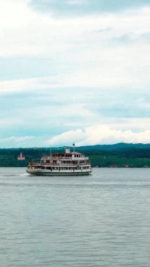 Yolcular su ve dağlara bakarken büyük bir tekne gölün karşısına geçiyor. Gökyüzü parçalı bulutlu ve bazı uzak binalar görülebilir..