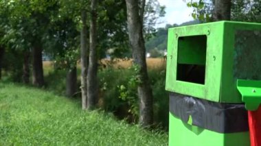 Yol kenarına uzun ağaçlar ve tarlalarla çevrili yeşil bir çöp kutusu konur. Güneş ışıl ışıl parlıyor. Manzarayı ve doğayı net bir şekilde görüyor..