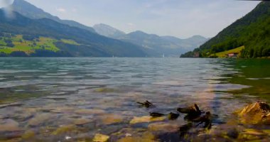 Parlak bir gökyüzünün altında verimli dağlarla çevrili berrak bir göl. Kayalık sahilde hafif dalgalar dolaşırken uzaktaki küçük tekneler görülebilir..