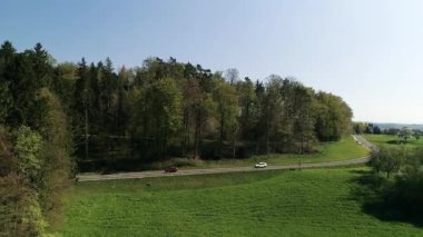 Arabalar yeşil tarlaları olan bir orman alanının yanındaki yol boyunca seyahat ederler. Güneşli bir gün. Yol kenarındaki ağaçlar gölge oluşturuyor..
