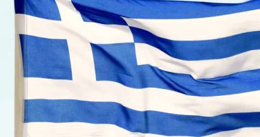 Yunanistan bayrağı açık mavi gökyüzünün altındaki tarihi bir alanda dalgalanırken görülüyor. Konumda bölgeden geleneksel mimari ve zengin tarih sergileniyor.