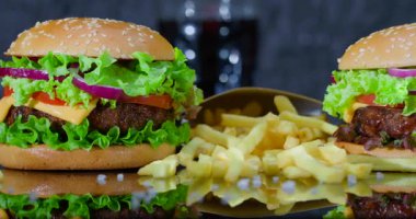 Hamburgerler marul, domates, soğan ve peynirle kaplı bir masanın üzerine konur. Altın patates kızartması servis ediliyor. Arka planda hafif bir içecek görülebilir..