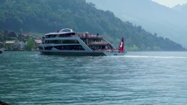 Bir tekne İsviçre 'de bir göl boyunca seyahat eder. İnsanlar gemide, sıcak güneşin ve açık gökyüzünün tadını çıkarıyorlar. Çevrede tepeler ve küçük binalar yer alıyor.
