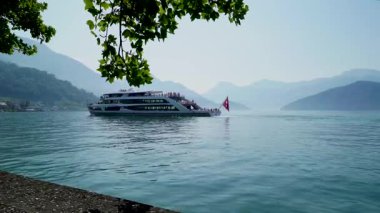 Arka planda dağlar olan bir göl boyunca bir tekne hareket ediyor. Güneş tepemizde parlarken insanlar kıyı şeridinden manzaranın tadını çıkarıyorlar. Su ışıkta parlıyor..