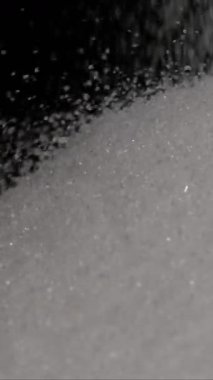 İnce beyaz şeker, mutfaktaki koyu bir yüzeye yavaşça dökülür. Işık taneciklerden yansıyarak, şeker yüzeyi kaplarken ışıltılı bir etki yaratıyor..