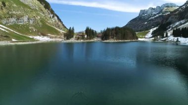Bir göl yeşil tepeler ve uzun ağaçlarla çevrilidir. Dağlar arka planda durur. Gökyüzü birkaç bulutla temiz. Bazı bölgelerde kar lekeleri kalıyor.