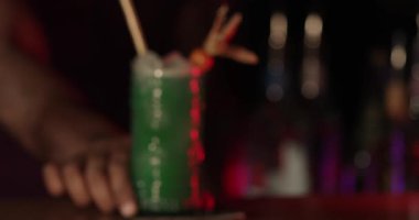 Bir barmen renkli bir içeceği garnitürle karıştırırken müşteriler de gece boyunca barda eğleniyor. İçkiler sergilendiği için atmosfer çok canlı..