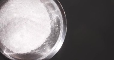 Bir insan kaseye şeker serpmek için ince bir elek kullanır. Mutfak yakındaki bir pencereden gelen doğal ışığa sahiptir. Odak noktası elek ve şeker..