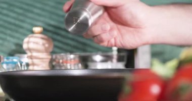 Bir kişi mutfakta yemek hazırlıyor. Taze sebzeler ve mutfak aletleriyle çevriliyken kızartma tavasına tuz ekliyorlar. Bu basit bir pişirme sürecini gösterir.