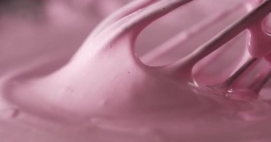 Bir insan, kapalı bir mutfakta çırpıcıyla pembe kremi kaseye karıştırır. Kremalı karışım pürüzsüz akıyor ve tatlı hazırlığı için hazır görünüyor..