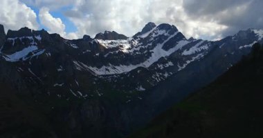 Mountains with snow-covered peaks rise against a cloudy sky. The green slopes below contrast with the rugged terrain of the national park. Sunlight touches the landscape.