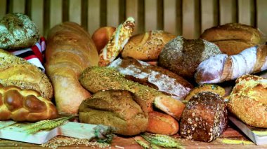 Tahta bir masanın üzerinde farklı türde ekmek vardır. Ekmekler arasında ekmekler, somunlar ve çeşitli tanelerden yapılan özel eşyalar var. Arka planda buğday var.