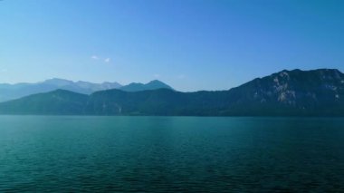 Açık mavi gökyüzü, güneş ışığı altında dağları ve sakin göl suyunu gösterir. Manzara bir yaz ortamında doğal güzelliği vurguluyor. Dağlar suyu yansıtır.