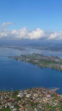 Mavi su üzerindeki kıyı kasabası, dağınık bulutlar ve uzak adalar, güneşli çatılar, marinalar ve botlar.