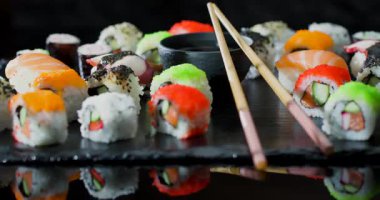 Parlak suşi ruloları siyah bir tabakta güzelce düzenlenir. Masada yemek çubukları ve küçük bir kase var. İnsanları yemeğin tadını çıkarmaya davet ediyorlar. Renkler öne çıkıyor ve dikkat çekiyor..