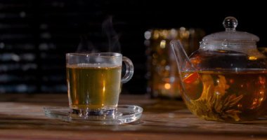 Bir bardak sıcak çay içerken buhar yükselir. Bir çaydanlık yanında daha fazla çayla oturuyor. Ortam mum ışığıyla sıcak, rahat bir ortam yaratıyor..