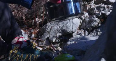 Bir kişi kamp sobasını kullanarak dağlarda yemek hazırlıyor. Yemek ekipmanları yerde, kayalar ve tırmanma ekipmanları görünür durumda. Güneş ışıldıyor..