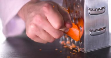 Bir insan metal rendesinde taze havuç rendeliyor. Rendelenmiş havuç karanlık bir yüzeye düşer ve öğle yemeğinde salata için basit malzemeleri hazırlama sürecini gösterir..