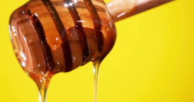 Tahta bir balıkçı balı damlarken tutar. Parlak sarı arka plan, balın yapışkan dokusunu ve altın rengini ön plana çıkarıyor. Damlama ve dökme eylemine odaklan.