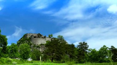 Yeşil ağaçlarla çevrili bir tepenin üzerinde bir kale bulunur. Gökyüzü bulutlarla mavidir. Sahne kırsal ve parlak, doğayı tam olarak gösteriyor..
