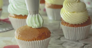 Bir kişi kapkekleri mutfakta değişik renklerde kremalarla süslüyor. Her kekin üstüne krema koymak için boru torbası kullanıyorlar..