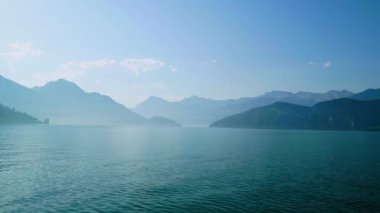 Açık mavi gökyüzü arka planda dağlar ve ön planda gölün sakin sularını gösteriyor. Güneş yüzeyde parlıyor, parlak bir görüntü yaratıyor..