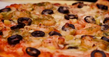 Bir şef üzerine karides ve siyah zeytin konmuş bir pizza hazırlar. Öğlen güneşi doğarken malzemeler yoğun bir mutfakta ayarlanır ve pişirilir..